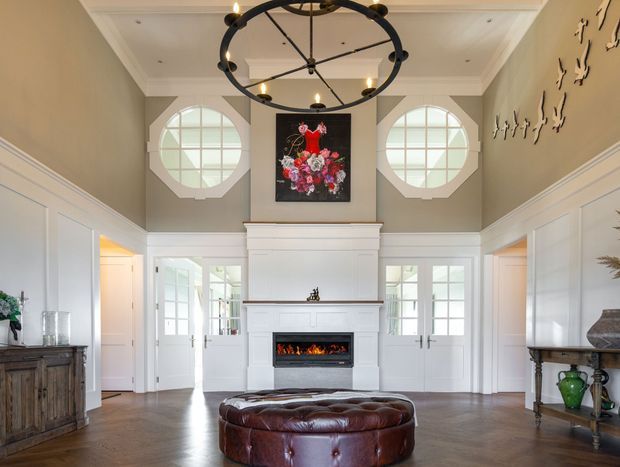 A bright, formal double-height living room centred on a white traditional fireplace with a built-in gas fire. The room features white wainscoting and panelled walls, symmetrical circular multi-pane windows, French doors, a large black circular chandelier, a dark timber floor, a tufted brown leather ottoman, and classic console furniture. Floral artwork and decorative wall pieces add visual interest.