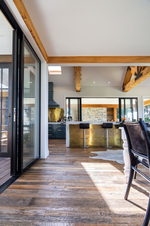 Bright open-plan kitchen and dining area viewed through large black-framed glazed doors. The space features warm timber flooring, exposed timber beams, a dark blue kitchen with brass-toned detailing, a substantial metallic island with bar stools, and a stone feature wall beyond. Leather dining chairs and strong natural light create a warm, rustic-contemporary atmosphere.
