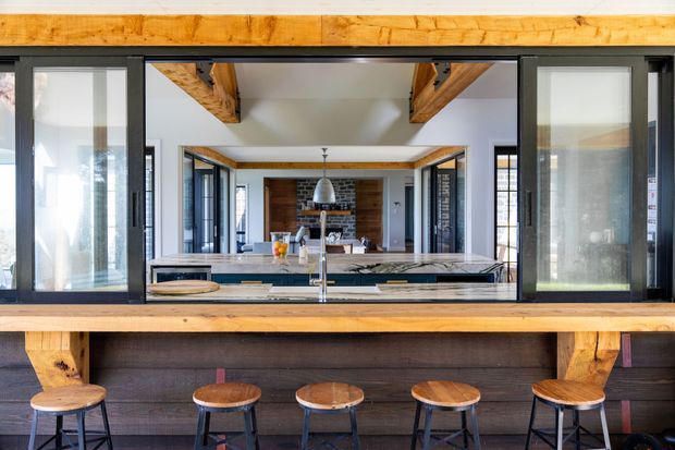 A contemporary kitchen viewed through wide black-framed glazed sliding doors, with a long dark island and timber benchtop, central sink and tap, and five timber-topped stools. Exposed timber beams and a timber-framed opening add rustic character, while the rear living area includes a dark brick feature wall, fireplace, and pendant light. The space is bright with a strong contrast between warm timber, dark cabinetry, black frames, and pale walls.