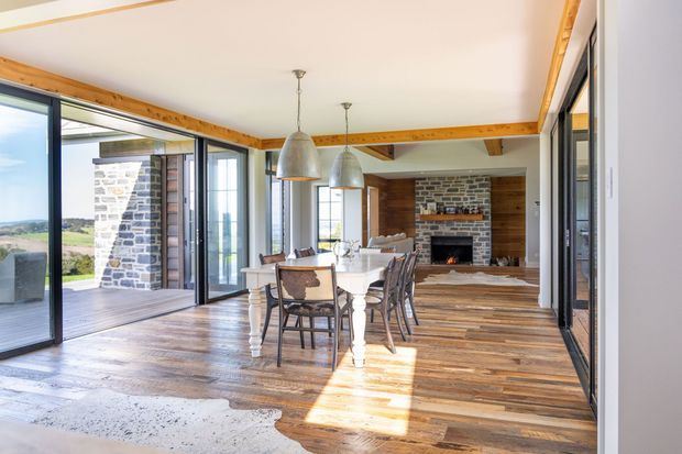 Bright open-plan dining and living area with extensive black-framed glazing, exposed timber beams and trim, wide-plank timber flooring, and a stone-and-brick fireplace with a wood-burning fire. A dining table sits centrally beneath two pendant lights, with a rural landscape visible through the folding glass doors.