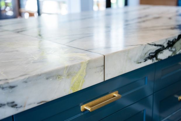 Close-up of a polished white natural stone kitchen benchtop with prominent grey and yellow veining, set above deep blue fixed cabinetry with brass-gold drawer handles. The image focuses on the material detail and joinery.