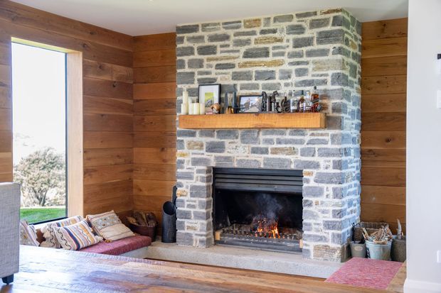 A rustic living room centred on a substantial natural stone fireplace with a timber mantel and lit fire. Horizontal timber wall panelling surrounds the fireplace, with a large window, built-in or low seating, patterned cushions, and timber flooring visible.