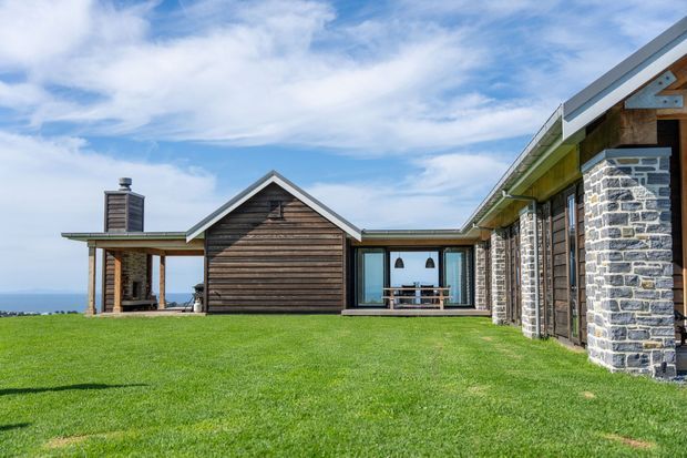 Exterior view of a low-slung multi-wing house set on a broad lawn, featuring dark horizontal timber cladding, substantial rough stone wall sections, exposed timber posts, large glazed openings, and a pitched gable roof. A covered outdoor area and dining setting are visible, with distant coastal or open landscape beyond the property.