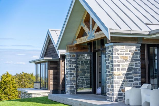 Exterior view of a contemporary alpine-style residential building with multiple steep gabled roofs, light metal roofing, dark timber wall cladding, and substantial grey stone masonry columns and walls. Large glazed openings and a covered entry terrace face a landscaped lawn, with white outdoor lounge chairs in the foreground.