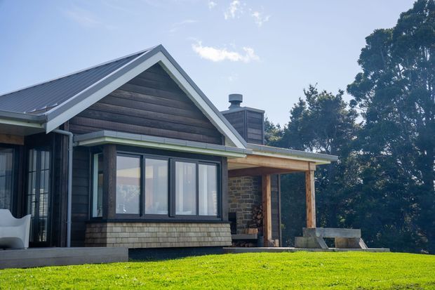 Exterior view of a contemporary gable-roofed home on a grassy site. The facade combines dark horizontal timber cladding, light trim, black-framed glazing, a stone masonry wall, and a covered timber porch or outdoor entertaining area. A chimney and stacked firewood are visible beside the porch.