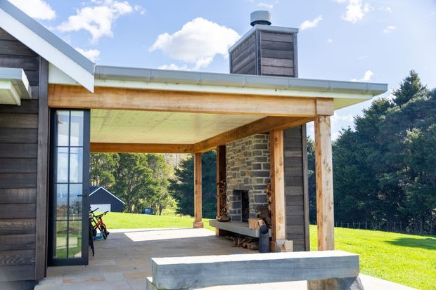 Exterior view of a contemporary timber-clad building with a broad covered outdoor entertaining area. The sheltered terrace features exposed timber posts and beams, a substantial stone fireplace and chimney, pale stone paving, and a built-in concrete or stone bench. Large glazed doors connect the terrace to the interior, with a sunny lawn and mature trees surrounding the structure.