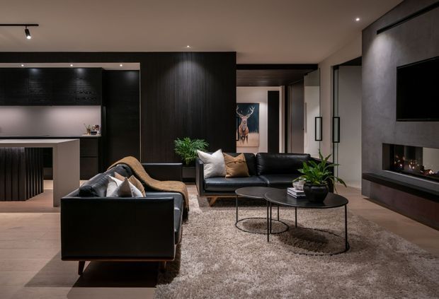 A dark, refined open-plan living room with a black leather sofa and armchair arranged around nested round coffee tables on a large textured rug. A contemporary fireplace and wall-mounted television occupy the right wall, while a compact kitchen with dark cabinetry and a pale island is visible behind the seating area. Recessed and track lighting, timber-look flooring, black built-in joinery, and decorative plants create a sophisticated, moody interior.