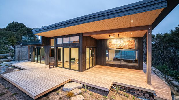 A contemporary single-storey timber-clad home with extensive floor-to-ceiling glazing, a broad flat roof with deep eaves, and a large timber deck extending around the facade. Warm timber soffits and dark structural framing create a sheltered outdoor living area, with rocky landscaped ground and dense vegetation surrounding the building.