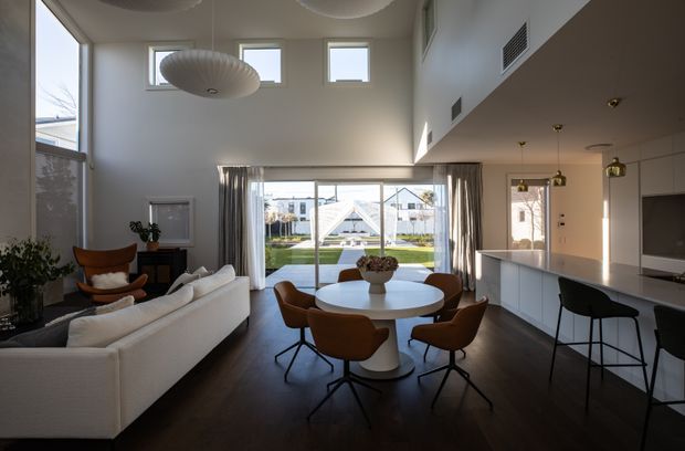 A bright, open-plan contemporary living, dining and kitchen space with a double-height ceiling and large glazed opening to the outdoor area. A white sectional sofa faces the dining zone, where a round white table is surrounded by brown upholstered chairs. The sleek white kitchen has a long island with dark bar stools, while sculptural pendant lights, dark timber flooring and full-height curtains add warmth and contrast.