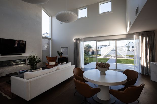 Bright contemporary open-plan living and dining area with a double-height ceiling, dark timber flooring, white walls, and large glazed doors opening to a landscaped outdoor area. A white sectional sofa faces a wall-mounted television and built-in media joinery, while a round white dining table with brown upholstered chairs sits beside the glazing. Floor-to-ceiling curtains and a sculptural pendant light complete the refined interior.