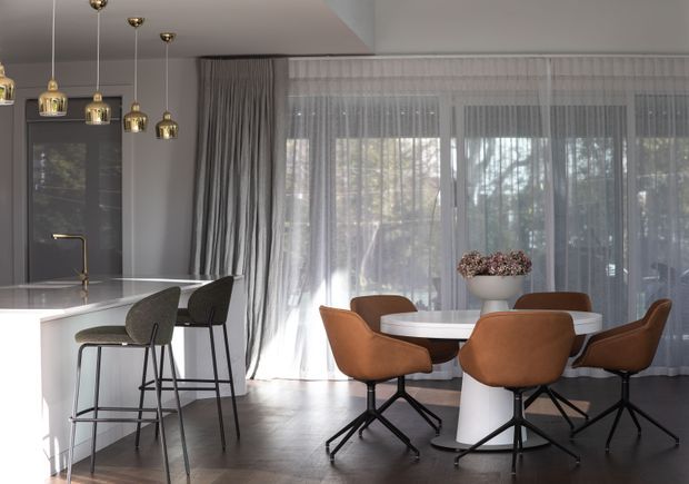 A refined open-plan kitchen and dining area with a bright white island, dark integrated cabinetry, and brass pendant lights. A round dining table with sculptural white pedestal base is surrounded by rust-brown upholstered chairs, while sheer curtains and grey drapery cover large windows overlooking trees. The space has dark timber flooring, a restrained neutral palette, and abundant filtered daylight.