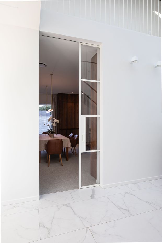 Bright interior view through a tall glazed pivot or hinged door into a dining room. The foreground has large white marble-look floor tiles and smooth white walls, with vertical white wall panelling and a high ceiling zone. The dining room beyond contains a timber dining table, upholstered brown leather dining chairs, dark timber cabinetry or panelling, pendant lighting, and a stair visible through the glazed door. The overall design is refined and contemporary with strong white and dark timber contrast.