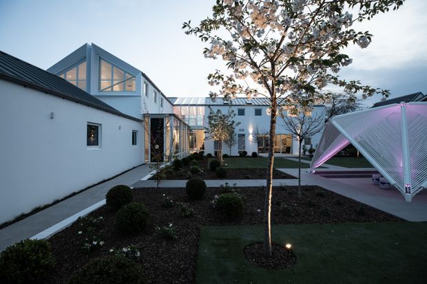 A landscaped contemporary courtyard between white rendered buildings, with dark pitched roofs, extensive glazed links and upper-level glazing. Geometric paths, clipped shrubs, lawn and a mature tree organize the courtyard. A striking angular outdoor pavilion or canopy with translucent ribbed surfaces is illuminated in pink-purple light on the right.