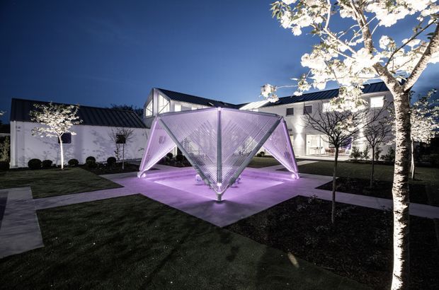Night-time exterior view of a landscaped courtyard or garden installation, featuring a sculptural, faceted translucent pavilion or canopy illuminated in purple. Pale concrete paths radiate through dark lawn and planted garden beds, with small trees and clipped shrubs. White contemporary buildings with dark metal pitched roofs surround the courtyard.