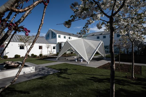 Exterior architectural view of a contemporary white building complex arranged around a landscaped courtyard. The buildings feature crisp geometric forms, white walls, dark metal standing-seam pitched roofs, and restrained rectangular windows. A striking folded, triangular metal canopy or pavilion occupies the paved central courtyard, surrounded by lawns, mature trees, garden beds, and concrete paths.