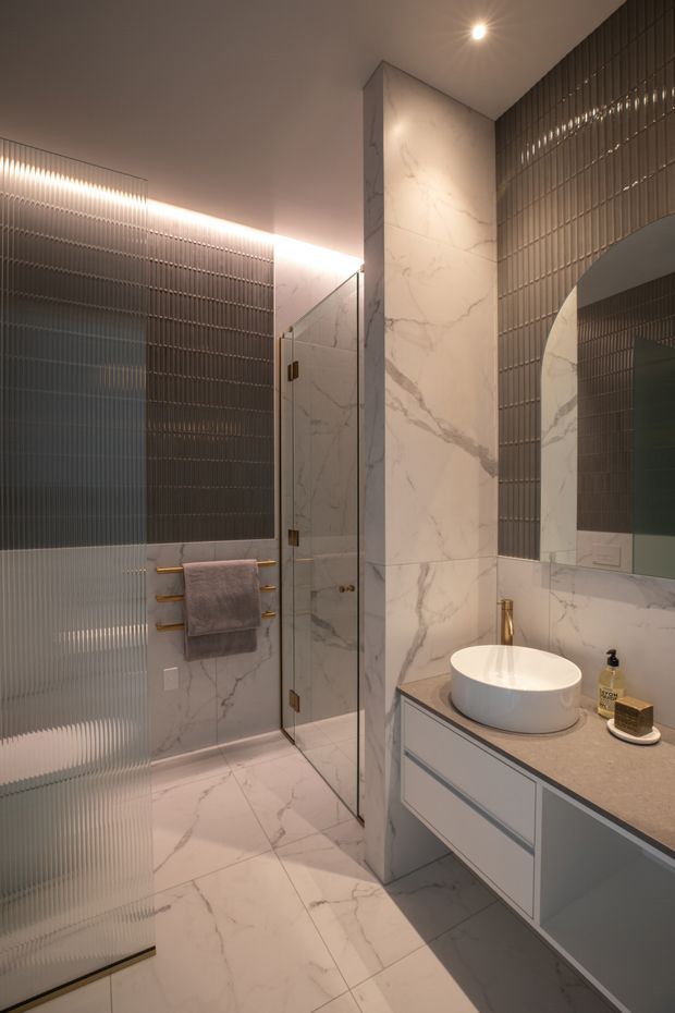 A compact, refined contemporary bathroom featuring white marble-look porcelain wall and floor surfaces, a grey floating vanity with a round vessel basin, frameless glass shower enclosure, large mirror, ribbed translucent glass screening, and warm integrated LED lighting. Brushed brass towel rails and tapware provide subtle accents against the muted grey and white palette.
