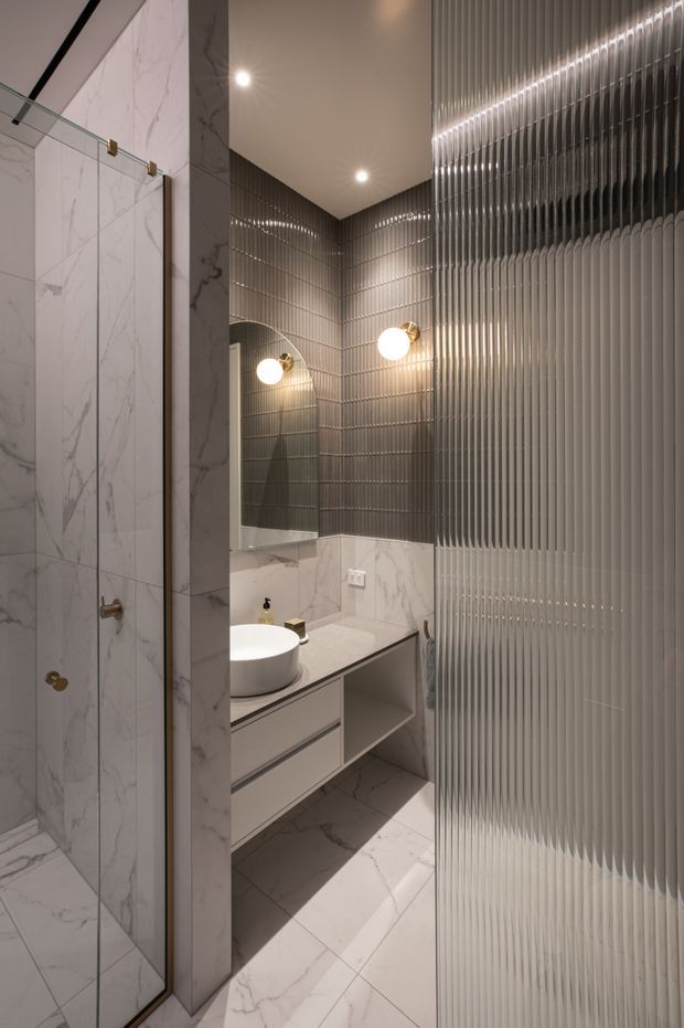 A compact, refined bathroom featuring white marble-look wall and floor tiles, a glass-enclosed shower, wall-mounted vanity with a round vessel basin, and a large mirror. Ribbed dark grey wall tiles create a textured feature surface, complemented by spherical wall lights and warm metallic fixtures. A fluted translucent glass or ribbed screen partially encloses the right side.