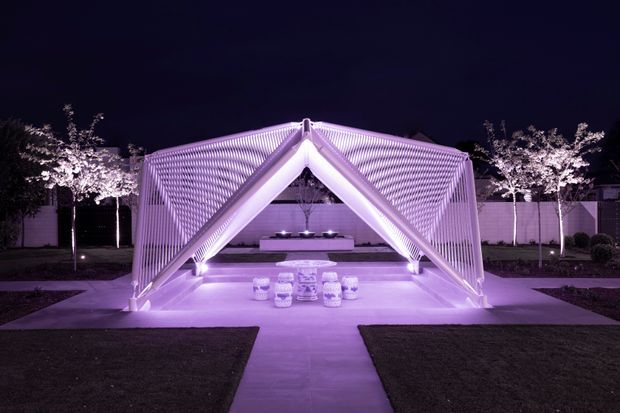 A symmetrical nighttime view of a sculptural outdoor pavilion or tent-like architectural structure, illuminated in vivid purple light. The structure has sharply pitched triangular framing, ribbed translucent or slatted side surfaces, and an open central entrance leading to a staged interior platform with low furnishings. A paved path approaches through manicured lawn, with small trees, hedges, and perimeter walls in the background.