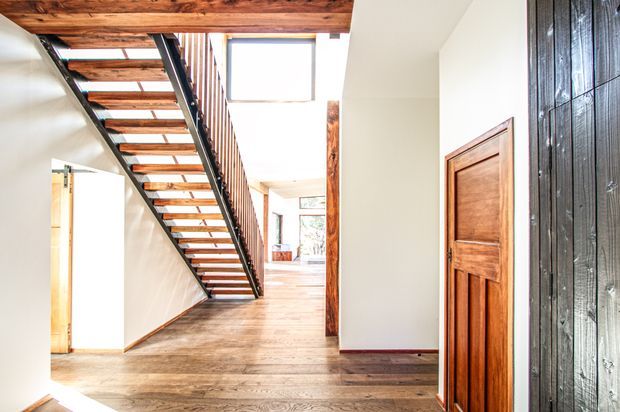 Bright interior hallway and landing beneath an open-riser timber-and-black-metal staircase. The space features warm timber flooring, exposed timber ceiling beams, white walls, a timber panelled door, and a dark vertically textured feature wall. Daylight enters through large upper-level glazing, creating an airy, double-height feel.