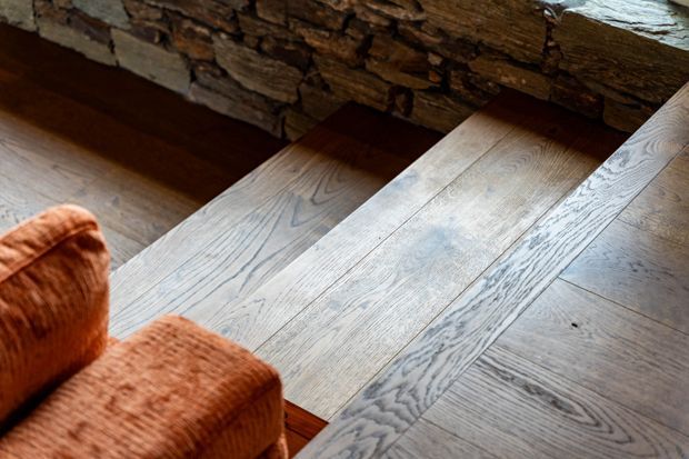 Close-up view of wide-plank timber flooring and matching timber stair treads beside a rustic stacked-stone wall. The flooring has a pale, weathered grain, while the partial upholstered armchair adds a warm rust-orange accent in the foreground.