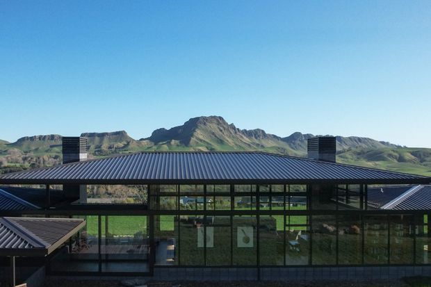 Exterior view of a contemporary, low-profile building with a broad dark metal pitched roof, extensive floor-to-ceiling glazing, and dark structural framing. Dramatic green volcanic or mountainous terrain forms the background, visible through and behind the building.