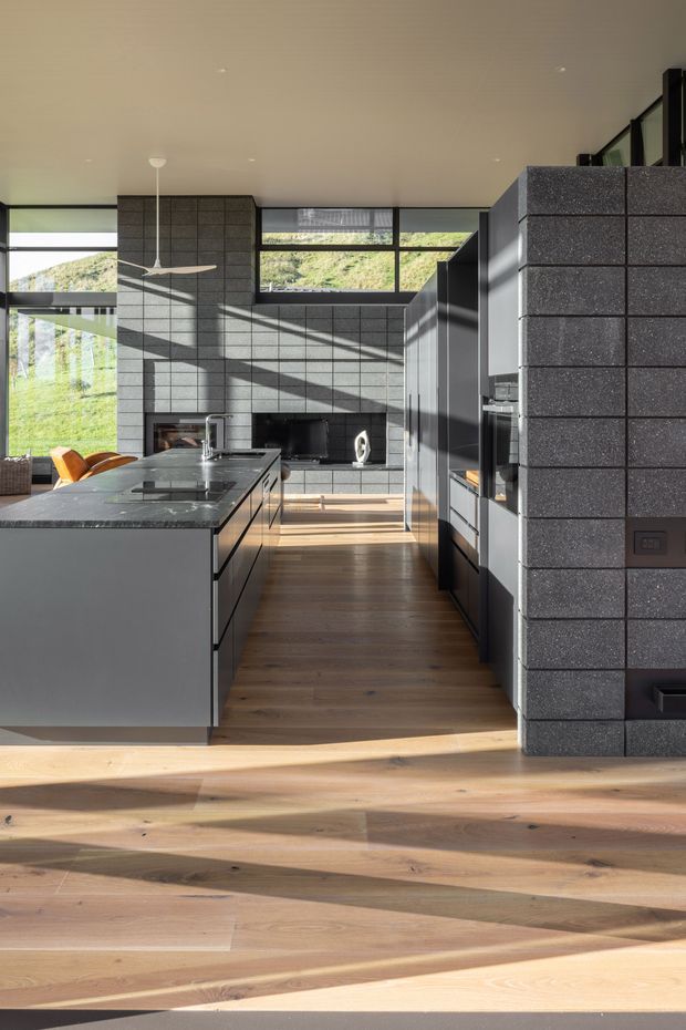 A contemporary open-plan kitchen with dark grey tiled masonry walls, charcoal built-in cabinetry and a large central island with a dark stone benchtop. Warm timber flooring extends through the room, while expansive black-framed glazing brings in strong sunlight and views of a green hillside. A ceiling fan, integrated appliances and a small adjoining seating area are visible.