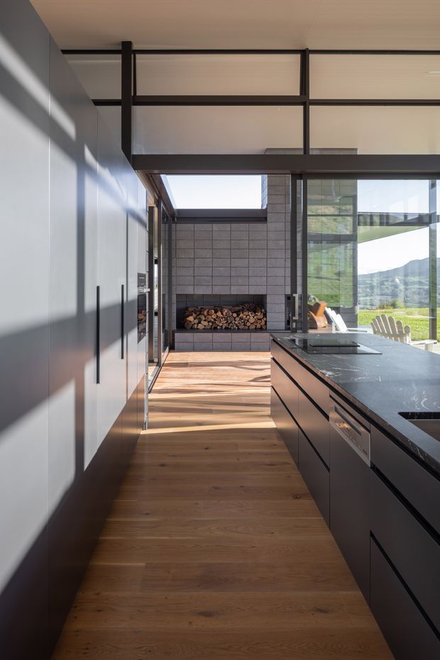 A contemporary galley kitchen with matte dark cabinetry on both sides, a dark stone island benchtop, warm timber flooring, and expansive black-framed glazing. At the far end is a tiled fireplace or wood-storage feature, while an outdoor landscape is visible through the glazing. A glazed upper-level void and exposed dark structural framing add architectural drama.