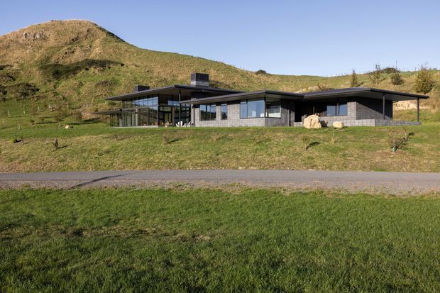 Low, single-storey contemporary house set into an expansive grassy hillside, with a broad flat roof, dark horizontal cladding, extensive floor-to-ceiling glazing, and a glazed corner living area. The rural sloped setting and surrounding hill dominate the exterior composition.
