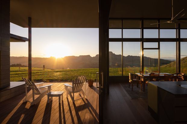 A warm sunset view from a contemporary open-plan home, showing a timber deck or covered terrace with outdoor chairs and stools on the left, and a dark timber-framed glazed dining area on the right. Floor-to-ceiling windows frame a sunlit rural valley and rugged mountain ridge. The interior includes a dining table and chairs, dark flooring, and a dark kitchen island or benchtop in the foreground.