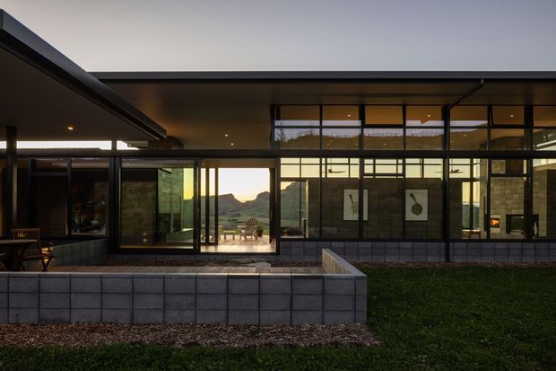 Low, single-storey modern residence photographed from the exterior at dusk. Broad flat roof planes and extensive dark-framed floor-to-ceiling glazing enclose a courtyard-like terrace, with exposed masonry walls and a raised paved planter or retaining wall in the foreground. Warm interior lighting contrasts with the shadowed facade, while distant mountains are visible through the glazing.