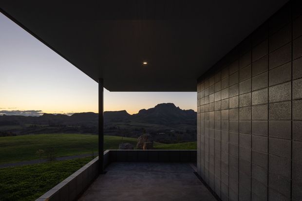 A minimalist covered outdoor terrace or balcony viewed at dusk, with a deep dark soffit, a slim structural post, concrete-paved floor, low parapet walls, and a textured modular masonry wall. The terrace frames a dramatic mountainous rural landscape beyond, with subdued natural light and strong geometric lines.