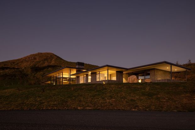 A single-storey contemporary home photographed at dusk, set into a grassy sloping site with a low, horizontally composed roofline. Warm interior lighting glows through extensive windows and glazed walls, while dark cladding and deep roof overhangs create a restrained modern silhouette against the hillside.