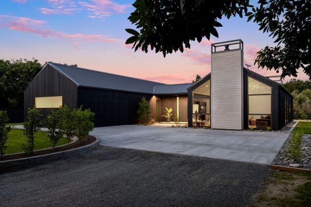 Exterior view of a contemporary single-storey house at dusk, composed of dark vertical cladding, pitched gabled roof forms, expansive glazed sections, and a prominent light-coloured chimney-like vertical volume. A broad concrete driveway and landscaped garden surround the building.