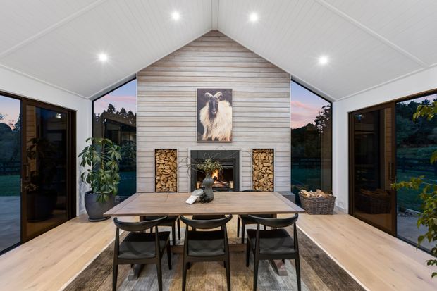 A bright, symmetrical open-plan dining and living space with a pitched white-lined ceiling and full-height glazing on both sides. A central feature wall clad in horizontal weathered timber boards incorporates a wood-burning fireplace, neatly stacked firewood niches, and a framed animal artwork. A timber dining table with dark chairs sits in the foreground, with pale timber flooring, a large neutral rug, indoor plants, and views of the surrounding landscape through the glass doors.