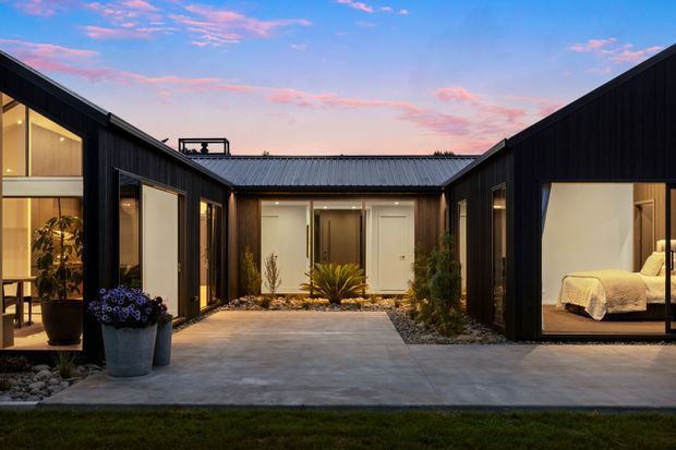 A modern single-storey residence arranged around a landscaped central courtyard at dusk. The dark vertical-clad wings form a U-shaped composition with low-pitched metal roofs, extensive floor-to-ceiling glazing, and warm interior lighting. A concrete-paved courtyard contains gravel beds and sculptural planting, while a bedroom is clearly visible through the glazed opening on the right and living or dining areas are visible on the left.