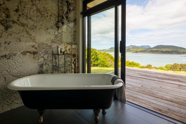 A luxurious freestanding clawfoot bathtub in a bathroom with dark flooring and a dramatic textured stone-look feature wall. Full-height black-framed glazing opens to a timber deck with expansive views across a calm bay and surrounding hills. The room combines an ornate vintage-style bath with a bold natural feature surface and a bright coastal outlook.