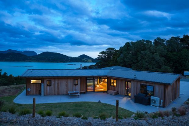Single-storey timber-clad house with a low-pitched metal roof, photographed at dusk from the landscaped foreground. Warm interior light glows through the windows and central glazing, while a large body of water, wooded hills, and dense trees form the backdrop. The building has a simple linear, contemporary composition with concrete paths and a small outdoor seating area.