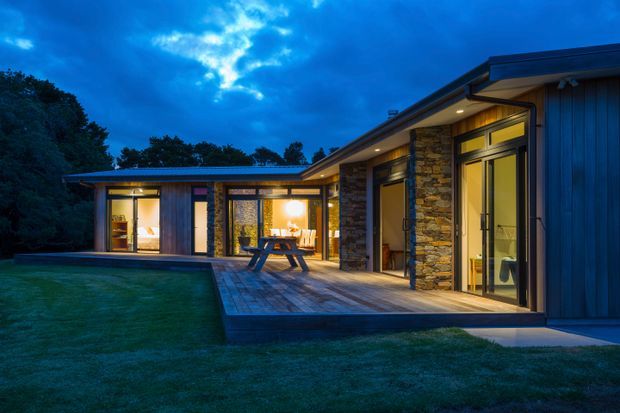 Exterior dusk view of a contemporary single-storey house with a low-pitched roof, dark vertical cladding, extensive floor-to-ceiling glazing, and substantial natural-stone wall sections. A timber deck and picnic-style outdoor table sit beside the illuminated interior, with lawn and mature trees surrounding the building.