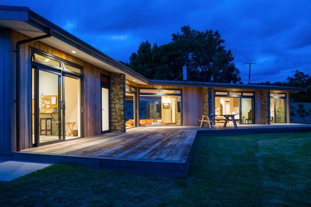 Twilight exterior view of a low, single-storey contemporary residence arranged around a broad timber deck. The home features extensive floor-to-ceiling glazing, dark-framed doors and windows, warm vertical timber wall cladding, and a natural stacked-stone feature wall. Interior lighting creates a warm glow, while a sheltered outdoor dining area and lawn are visible in the foreground.