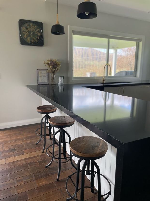 A contemporary kitchen with a long black stone-look island and integrated breakfast bar, lined with three industrial-style stools featuring dark metal frames and round timber seats. White vertical-panel cabinetry sits beneath the island, with matching dark benchtops continuing around the kitchen. A large window above the sink brings in natural light and overlooks trees, while black pendant lights, timber flooring, and a decorative wall clock complete the space.