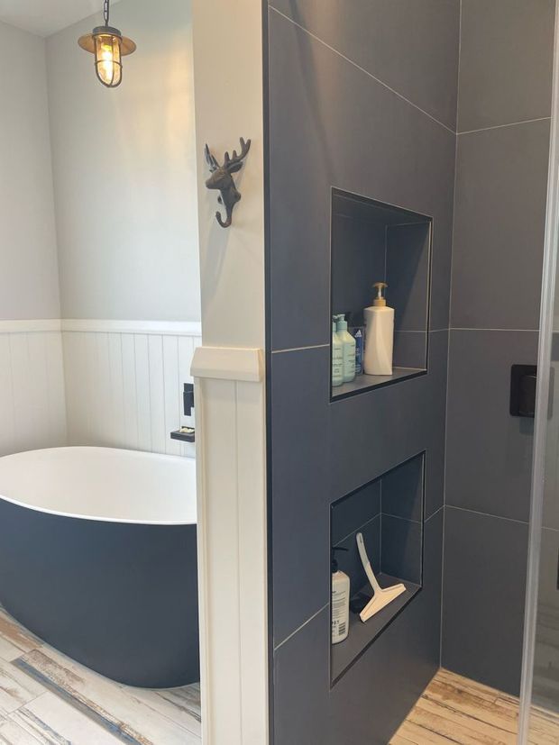 A compact, stylish bathroom featuring a freestanding dark-sided bathtub beside white vertical wall panelling, a dark grey tiled shower wall with two recessed storage niches, and a glass shower enclosure. A rustic industrial pendant light and decorative deer-head wall hook add character. Timber-look flooring and crisp white trim create contrast with the dark finishes.