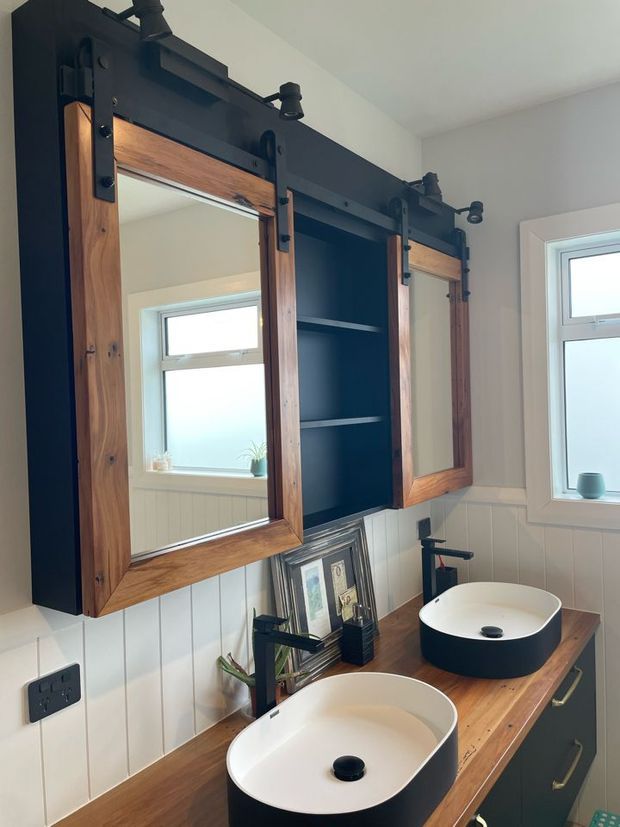 A bright bathroom with a long timber vanity featuring two white vessel basins, black taps, dark cabinetry, and a large central open shelf. Above the vanity are matching timber-framed mirrored cabinets with black barn-door-style hardware and integrated spotlights. White vertical wall panelling, black fixtures, and warm timber surfaces create a rustic-industrial character.