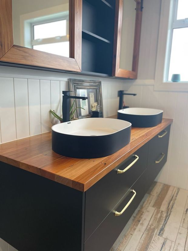 A compact, warmly styled bathroom vanity with two matte dark vessel basins set into a solid timber benchtop. The room features dark cabinetry with brass handles, white vertical wall panelling, timber-framed mirrored cabinetry, black tapware, and a light wood-look floor.