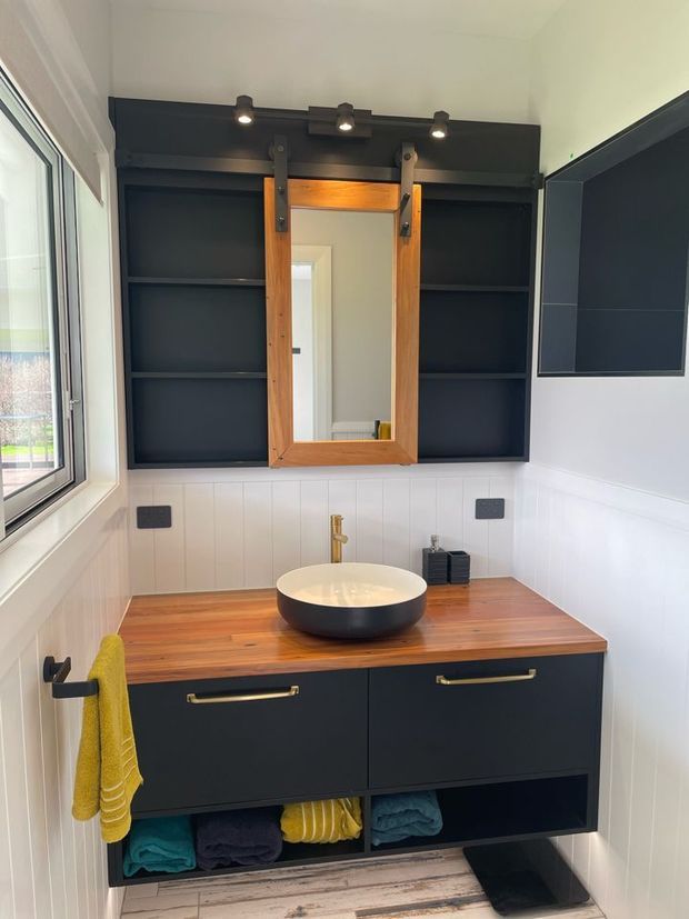 A compact, contemporary bathroom vanity with a dark charcoal built-in cabinet, warm timber benchtop, vessel basin and brass tapware. Above the vanity are open dark shelving units framing a timber-framed mirror, with integrated spot lighting. White vertical wall panelling, a large window and understated black hardware complete the space.