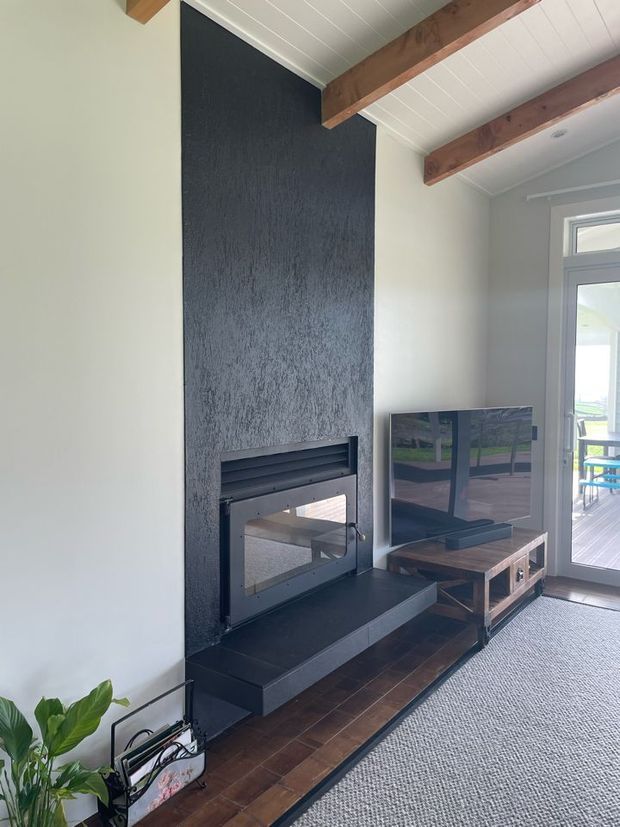 A contemporary living room featuring a tall dark textured fireplace surround with a built-in glass-front fireplace and black hearth. Exposed timber beams span a white-lined ceiling, while a timber television console sits beside the fireplace. Glazed doors open to a timber deck, and a large neutral rug covers part of the dark timber floor.