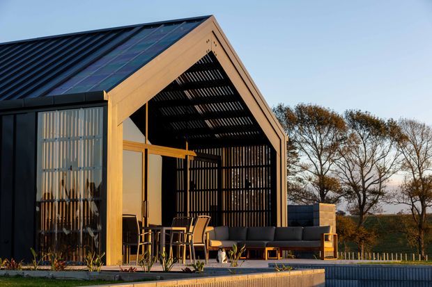 Exterior view of a contemporary gable-roof pavilion or house, featuring dark metal cladding, warm timber framing, a large open slatted facade, glazed sliding doors, and an outdoor entertaining area with dining and lounge furniture. The building sits beside a raised pool or terrace edge with landscaped surroundings.