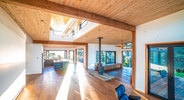 Bright open-plan interior with extensive warm timber lining across the ceiling, exposed timber beams, polished timber flooring, white walls, large windows and sliding glass doors opening to a deck and leafy outlook. A central wood-burning fireplace and sunlit living area are visible, with a partial kitchen or dining zone beyond.