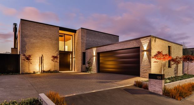 Twilight exterior view of a contemporary multi-volume brick residence. The home features warm architectural lighting, a recessed glazed entry, a broad dark garage door, paved driveway, and landscaped frontage with ornamental grasses and a small tree.