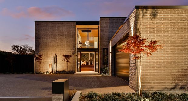 Twilight exterior view of a contemporary brick residence with a low, rectilinear composition. The entry is framed by warm floor-to-ceiling glazing, while a recessed garage sits to the right behind a projecting brick wing. Paved forecourt, dark fencing, low planting, and a sculptural Japanese maple complete the restrained architectural presentation.