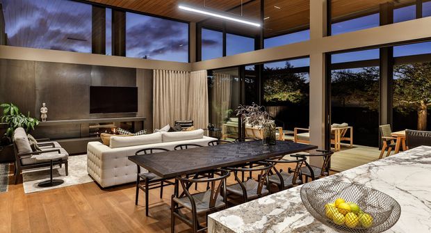 A sophisticated open-plan living and dining area with floor-to-ceiling glazing, a dark stone feature wall and television zone, cream sofas, a long dark dining table, and a marble-look kitchen island in the foreground. Warm timber flooring and a timber-lined ceiling contrast with the dark metal window frames, while evening light and garden views create an inviting atmosphere.